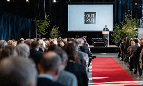 Speaker on stage in front of an OUTPUT screen, with the audience in the foreground