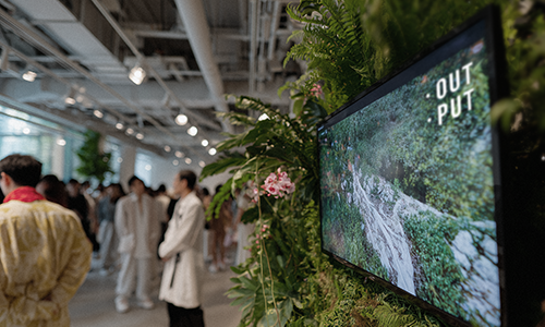 Visitors in the exhibition hall beside an OUTPUT display screen and plants