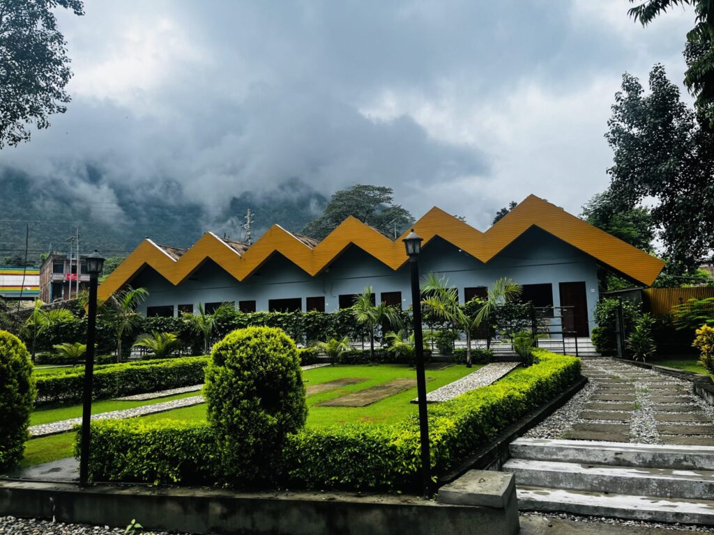 Low blue-grey building with an orange zigzag roofline behind trimmed hedges, a lawn and a stone path, with forested hills and low cloud behind