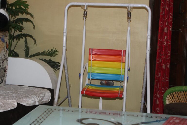 Rainbow-striped baby swing in a white metal frame beside a glass-topped table in a living room, with a red curtain and a potted plant