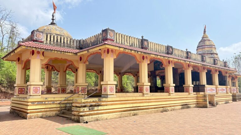 Open pavilion with a row of painted columns and arched bays under a tiled roof with a dome, against a bright blue sky
