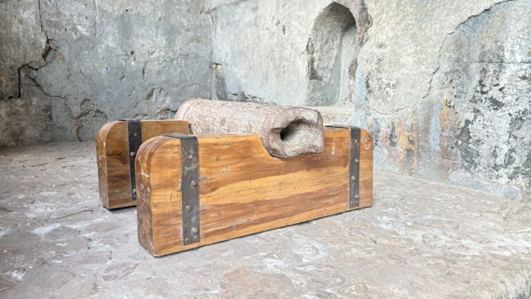 Two wooden blocks with iron bands and a carved wooden piece resting on a stone ledge in front of a weathered wall