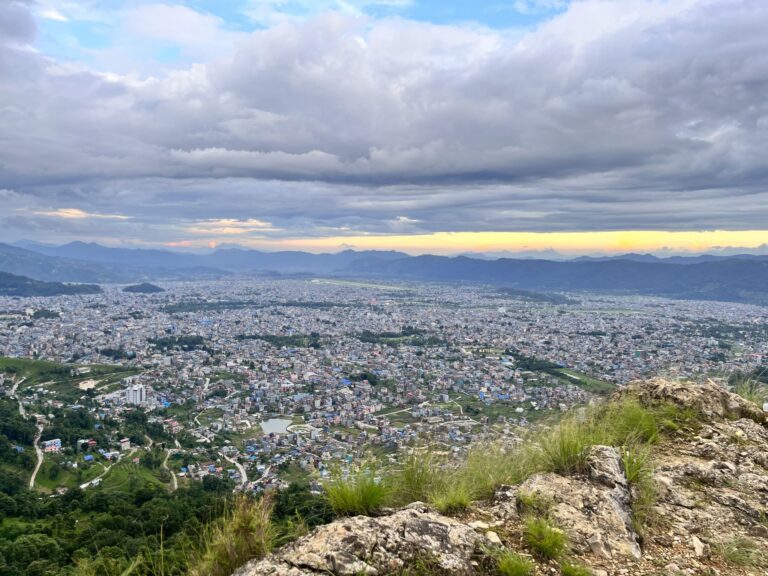 Wide view over a city at dusk beneath heavy clouds with a band of golden light at the horizon